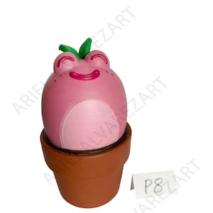 May include: A pink frog figurine with a smiling face and green leaves on its head, sitting in a brown flower pot. The frog has a lighter pink belly. A small white tag with the text "P8" is visible.