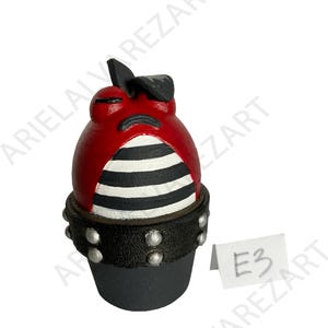 May include: A red and black figurine with a striped midsection and a studded base. The figurine has a small black hat and a tag labeled "E3".