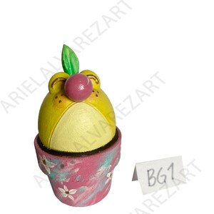May include: A whimsical, hand-painted ceramic figurine of a yellow creature with a pink nose and green leaf, sitting in a pink and blue flower pot. The pot is decorated with white flowers. The tag reads "BG1".