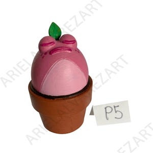 May include: A whimsical, hand-painted frog figurine in a brown flower pot. The frog is primarily pink with a lighter pink belly and closed eyes. A green leaf sits atop its head. A small white tag with "P5" is visible.