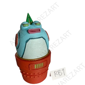 May include: A whimsical, hand-painted figurine of a robot frog. The frog has a light blue head with orange accents, a pink mouth, and a green triangle on top. It sits atop a red, cylindrical base.