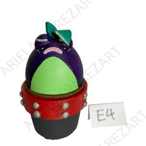 May include: A whimsical, hand-painted figurine of a frog-like creature. The figure features a purple head, green belly, and a red collar with silver studs. The base is black, and the frog has a green leaf on its head. A small tag reads "E4".