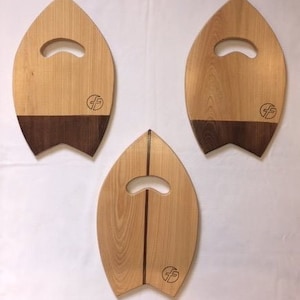 May include: Three wooden cutting boards shaped like surfboards. Each board has a handle and a dark wood inlay. The boards are all natural wood color and have a logo on the bottom.