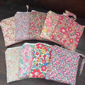 May include: A collection of nine rectangular fabric pouches featuring various floral patterns in shades of pink, red, yellow, green, and blue. Each pouch is quilted, includes a white zip closure, and has a small fabric tab attached to the side.