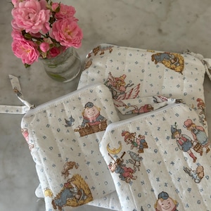 Puede incluir: Tres bolsas de tela blanca con un estampado infantil caprichoso con personajes, letras y números. Las bolsas tienen cierre de cremallera y un pequeño lazo de tela. Un pequeño jarrón de cristal contiene rosas rosas.