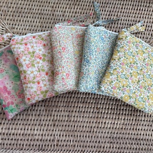 May include: Five floral print zippered pouches in various colors: pink, green, blue, and yellow. Each pouch has a matching fabric pull tab. The pouches are arranged on a woven wicker surface.