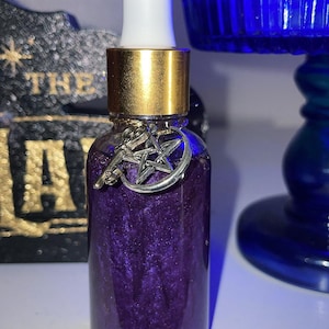 May include: A small, clear glass bottle filled with a shimmering purple liquid. The bottle has a gold-colored cap and a white dropper. A silver charm with a star and crescent moon hangs from the bottle. The background includes the text "THE LAY".