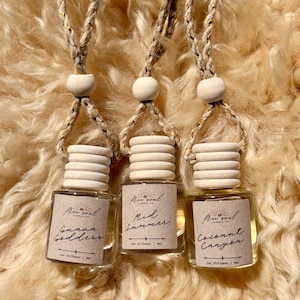 May include: Three car air fresheners with wooden tops and hanging cords. Each glass bottle has a label with the words "Guava Goddess", "Mid Summer", and "Coconut Canyon". The background is a fuzzy, light brown material.