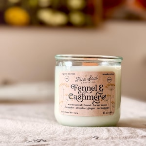 May include: A clear glass jar candle with a label that reads "Fennel & Cashmere" from Free Soul Candle Co. The label lists ingredients like warm santal, fennel, and ginger. The candle is 10 oz (283 g) and has a wooden wick.