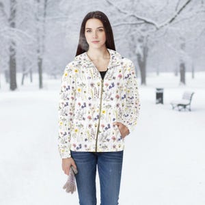 May include: A white zip-up jacket featuring a colourful botanical print of various wildflowers, including yellow, purple, and pink blossoms. The jacket has long sleeves, a high collar, and a central zipper closure, worn over a black top with blue jeans.