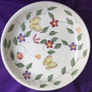 Könnte beinhalten: Weiße Keramik-Servierschale mit Blumenmuster. Die Schale zeigt grüne Ranken, gelbe Schmetterlinge, rote und violette Blumen und einen Marienkäfer.