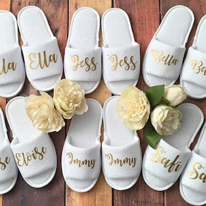 May include: Eight pairs of white slippers with gold lettering. The names Ella, Jess, Bella, Eloise, Jimmy, and Safi are written on the slippers.