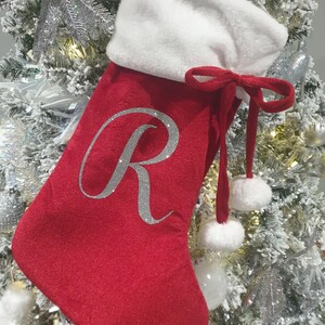 Puede incluir: Calcetín navideño rojo con puño blanco y letra "R" plateada con purpurina. El calcetín tiene un lazo rojo y dos pompones blancos. El calcetín está colgado de un árbol de Navidad decorado.