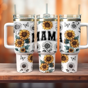 May include: White stainless steel tumbler with a black and gold lid and a black handle. The tumbler has a sunflower and butterfly design and the word "MAMA" printed on the side.