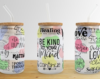 Op de afbeelding: Drie glazen bekers met houten deksels en heldere rietjes. Elke beker heeft een witte achtergrond met kleurrijke abstracte vormen en inspirerende tekst in het zwart. De tekst op de bekers is "Be a Human", "Be Kind to Your Mind" en "Self Love".
