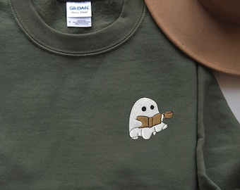 May include: Olive green sweatshirt with an embroidered ghost reading a book and holding a cup. The sweatshirt is paired with blue jeans, a brown hat, and a coffee cup. The sweatshirt has a crew neck and long sleeves.