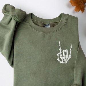 Puede incluir: Una sudadera verde con una mano de esqueleto bordada en blanco haciendo el gesto de rock and roll.