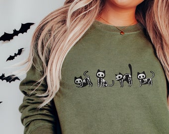 Puede incluir: Una sudadera verde con un gráfico de cinco gatos esqueleto en fila. Los gatos son negros con huesos blancos. La sudadera la lleva puesta una persona que lleva pantalones vaqueros azules.