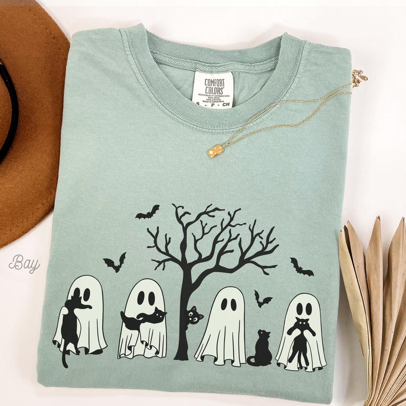 Puede incluir: Una camiseta azul claro con un gr&aacute;fico negro de cuatro fantasmas sosteniendo gatos negros frente a un &aacute;rbol desnudo con murci&eacute;lagos volando alrededor. El texto "COMFORT COLORS" es visible en la etiqueta.