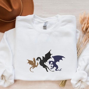 May include: White sweatshirt featuring a black, blue and gold silhouette of three dragons.