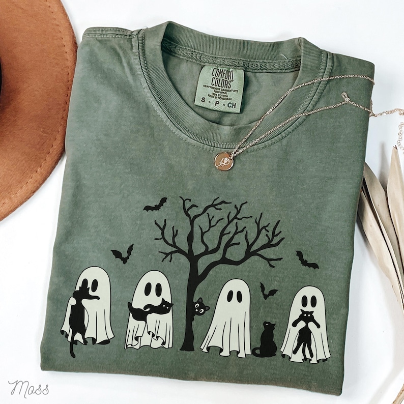 Puede incluir: Camiseta verde con un gr&aacute;fico en blanco y negro de fantasmas sosteniendo gatos negros frente a un &aacute;rbol desnudo. El texto "COMFORT COLORS" es visible en la etiqueta.