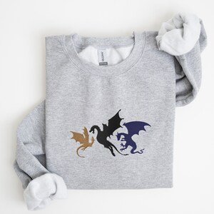 May include: A grey sweatshirt featuring a design of three dragons in black, gold, and blue. The dragons are facing each other with their wings outstretched.