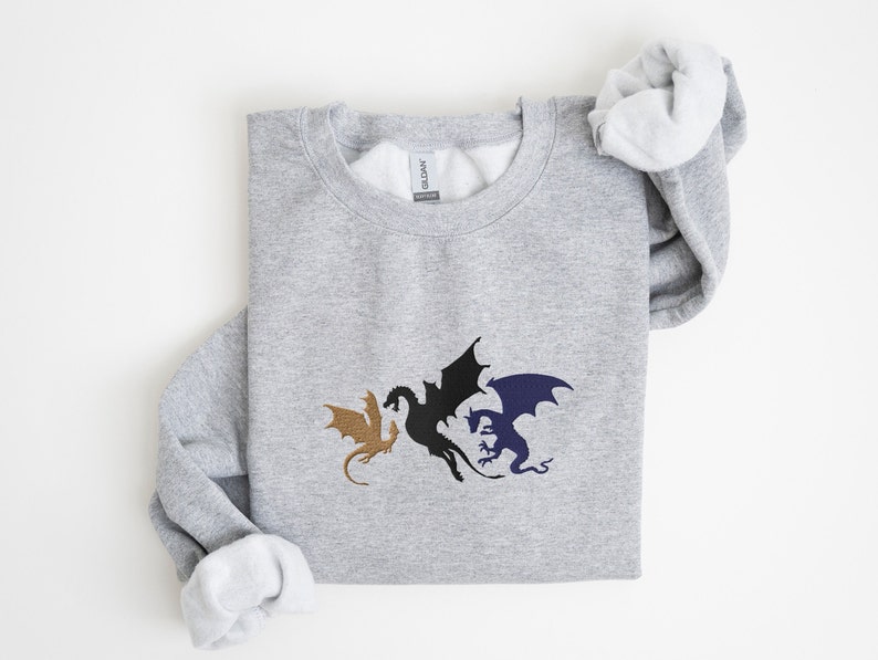 May include: A grey sweatshirt featuring a design of three dragons in black, gold, and blue. The dragons are facing each other with their wings outstretched.