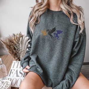 May include: A dark grey sweatshirt featuring a graphic of three dragons in flight. The dragons are black, gold, and blue.