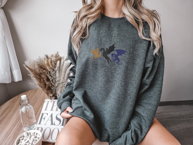 May include: A dark grey sweatshirt featuring a graphic of three dragons in flight. The dragons are black, gold, and blue.