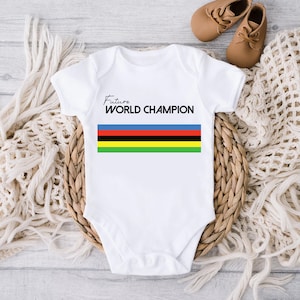 K&ouml;nnte beinhalten: Ein wei&szlig;er kurz&auml;rmeliger Babybody mit dem Schriftzug Future WORLD CHAMPION in schwarzen Buchstaben, platziert &uuml;ber f&uuml;nf horizontalen Streifen in den Farben Blau, Rot, Schwarz, Gelb und Gr&uuml;n, zentriert auf der Vorderseite des Kleidungsst&uuml;cks.