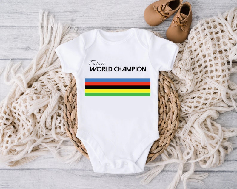 K&ouml;nnte beinhalten: Ein wei&szlig;er kurz&auml;rmeliger Babybody mit dem Schriftzug Future WORLD CHAMPION in schwarzen Buchstaben, platziert &uuml;ber f&uuml;nf horizontalen Streifen in den Farben Blau, Rot, Schwarz, Gelb und Gr&uuml;n, zentriert auf der Vorderseite des Kleidungsst&uuml;cks.