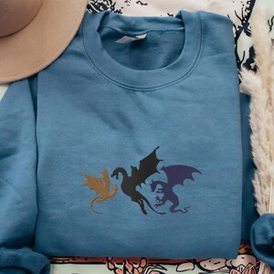 May include: A blue sweatshirt featuring a graphic of three black dragons with purple and brown accents.
