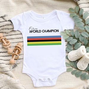 K&ouml;nnte beinhalten: Ein wei&szlig;er kurz&auml;rmeliger Babybody mit dem schwarzen Schriftzug Future WORLD CHAMPION &uuml;ber f&uuml;nf horizontalen Streifen in Blau, Rot, Schwarz, Gelb und Gr&uuml;n. Das Kleidungsst&uuml;ck hat einen Rundhalsausschnitt und drei Druckkn&ouml;pfe im Schrittbereich.