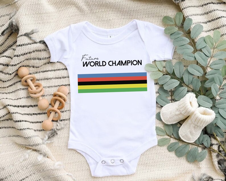 K&ouml;nnte beinhalten: Ein wei&szlig;er kurz&auml;rmeliger Babybody mit dem schwarzen Schriftzug Future WORLD CHAMPION &uuml;ber f&uuml;nf horizontalen Streifen in Blau, Rot, Schwarz, Gelb und Gr&uuml;n. Das Kleidungsst&uuml;ck hat einen Rundhalsausschnitt und drei Druckkn&ouml;pfe im Schrittbereich.