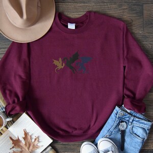 May include: Burgundy sweatshirt featuring a black, gold, and blue dragon design. The sweatshirt is laid flat on a wooden surface with a hat, book, and jeans nearby.