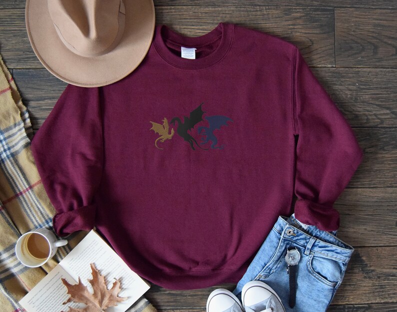 May include: Burgundy sweatshirt featuring a black, gold, and blue dragon design. The sweatshirt is laid flat on a wooden surface with a hat, book, and jeans nearby.