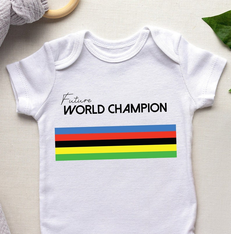 K&ouml;nnte beinhalten: Ein wei&szlig;er kurz&auml;rmeliger Babybody mit dem schwarzen Schriftzug Future WORLD CHAMPION &uuml;ber einem horizontalen Streifenmuster in den Farben Blau, Rot, Schwarz, Gelb und Gr&uuml;n, das mittig auf der Brust aufgedruckt ist.