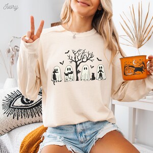 Puede incluir: Una sudadera de manga larga de color crema con un estampado gr&aacute;fico de cinco fantasmas y cuatro gatos negros. Los fantasmas llevan sombreros y bufandas. Los gatos est&aacute;n sentados delante de los fantasmas. Hay un &aacute;rbol detr&aacute;s de los fantasmas. La sudadera tiene cuello redondo.
