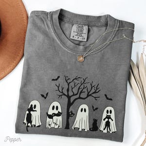 Puede incluir: Una camiseta gris con un gr&aacute;fico en blanco y negro de fantasmas sosteniendo gatos frente a un &aacute;rbol con murci&eacute;lagos volando alrededor. La etiqueta dice "COMFORT COLORS".