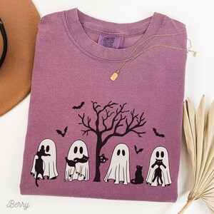 Puede incluir: Una camiseta morada con un gr&aacute;fico de cuatro fantasmas sosteniendo gatos negros frente a un &aacute;rbol desnudo. El texto "Comfort Colors" es visible en la camiseta.