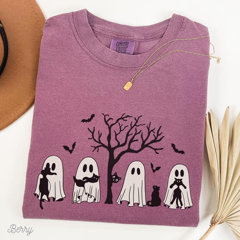 Puede incluir: Una camiseta morada con un gr&aacute;fico de cuatro fantasmas sosteniendo gatos negros frente a un &aacute;rbol desnudo. El texto "Comfort Colors" es visible en la camiseta.