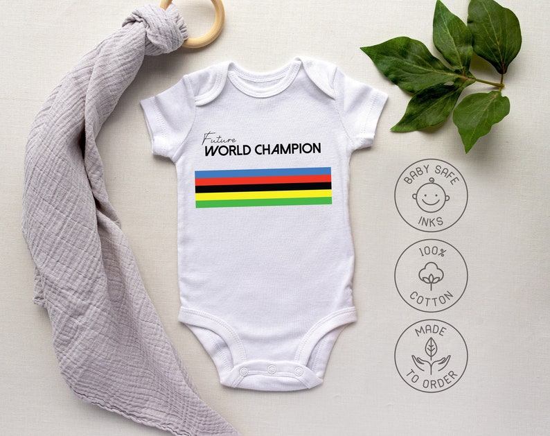 K&ouml;nnte beinhalten: Ein wei&szlig;er kurz&auml;rmeliger Babybody mit dem Text Future WORLD CHAMPION in schwarzer Schrift &uuml;ber einem horizontalen Streifenmuster in Blau, Rot, Schwarz, Gelb und Gr&uuml;n. Drei kreisf&ouml;rmige Symbole zeigen babyfreundliche Tinte, 100% Baumwolle und auf Bestellung gefertigt.