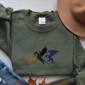 May include: A green sweatshirt with a three-headed dragon design embroidered on the front. The dragons are gold, black, and purple.