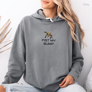 Op de afbeelding: Een grijze hoodie met een geborduurde afbeelding van een goudkleurige krabklauw boven de zwarte tekst FIST MY BUMP. Het kledingstuk is voorzien van een capuchon met witte trekkoorden en lange mouwen met geribbelde boorden aan de uiteinden.