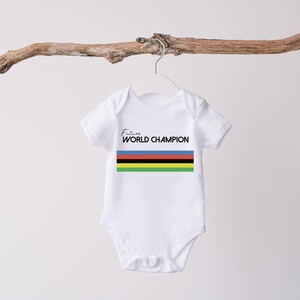 K&ouml;nnte beinhalten: Ein wei&szlig;er kurz&auml;rmeliger Babybody mit dem Schriftzug Future WORLD CHAMPION in schwarzen Buchstaben &uuml;ber einem horizontalen Streifenmuster in den Farben Blau, Rot, Schwarz, Gelb und Gr&uuml;n. Das Kleidungsst&uuml;ck hat einen Rundhalsausschnitt und Druckkn&ouml;pfe.