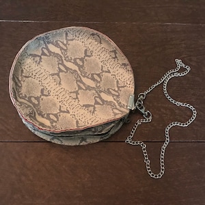May include: A round, light brown snakeskin-patterned purse with a silver chain strap.