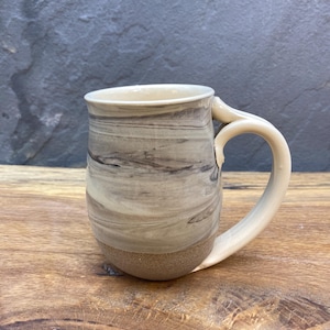 May include: A ceramic mug with a light brown and gray marbled design. The mug has a rounded body and a handle.
