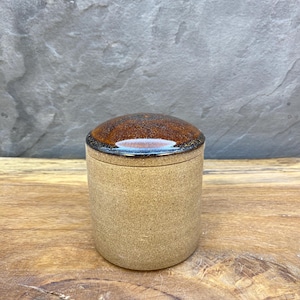 May include: A small, cylindrical ceramic jar with a lid. The jar is a light tan color with a textured surface. The lid is a glossy brown with a dark brown rim. The jar is sitting on a wooden surface.