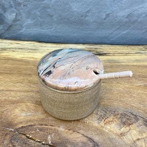 May include: A ceramic salt cellar with a matching lid and small spoon. The lid features a marbled pattern in shades of pink, blue, and brown. The container and spoon are a light tan color. The salt cellar is on a wooden surface.