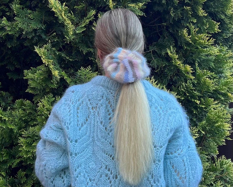 May include: A woman with long blonde hair wearing a light blue knitted sweater with a large open stitch pattern. Her hair is pulled back in a ponytail and secured with a colorful fuzzy scrunchie.